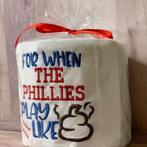 May include: A white toilet paper roll with red, blue, and brown embroidery. The text reads "For when the Phillies play like" with a brown poop emoji.