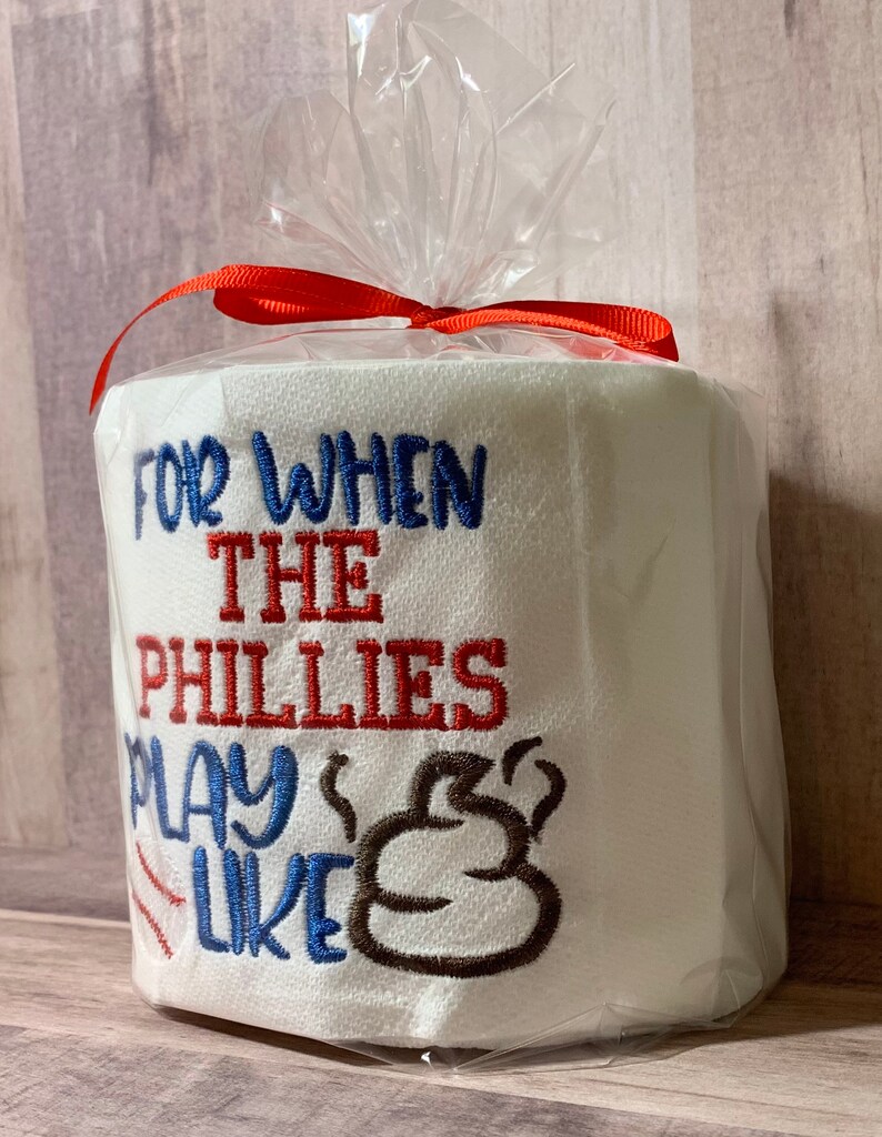 May include: A white toilet paper roll with red, blue, and brown embroidery. The text reads "For when the Phillies play like" with a brown poop emoji.