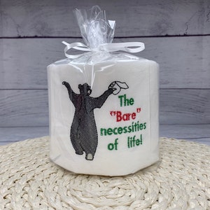 May include: A white toilet paper roll with an embroidered design of a black bear holding a roll of toilet paper. The text reads "The "Bare" necessities of life!"