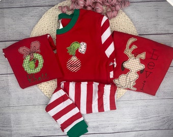 May include: Red and white striped pajamas with a green trim. The top has a green and white wreath with a bow, a green Grinch holding a red and white ornament, and a brown dog with antlers. The pajamas are personalized with the names "Rhett" and "Levi".