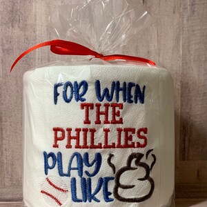 May include: A white toilet paper roll with red, blue, and brown embroidery that reads "For when the Phillies play like" with a baseball and a poop emoji. The roll is wrapped in clear plastic with a red ribbon.