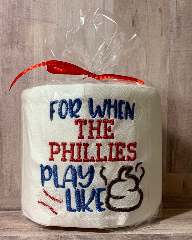 May include: A white toilet paper roll with red, blue, and brown embroidery that reads "For when the Phillies play like" with a baseball and a poop emoji. The roll is wrapped in clear plastic with a red ribbon.