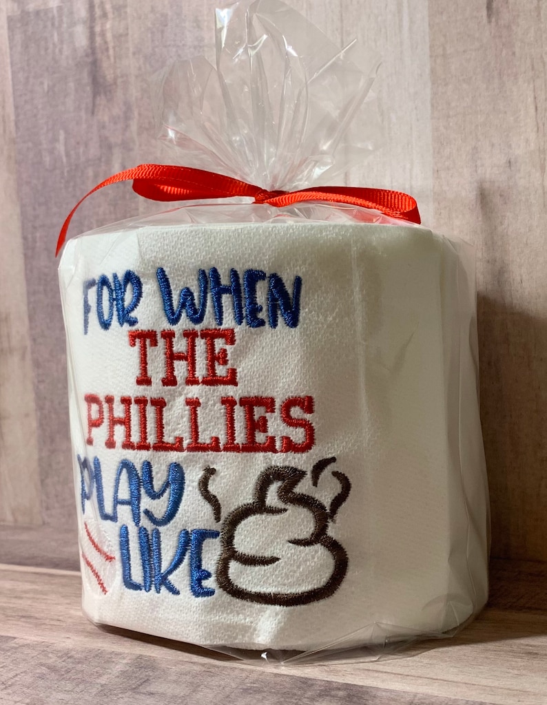 May include: A white toilet paper roll with red, blue, and brown embroidery. The text reads "For when the Phillies play like" with a brown poop emoji.