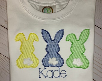 May include: White t-shirt with three embroidered bunny silhouettes in yellow, blue, and green. The name "Kade" is embroidered in blue below the bunnies. The shirt has a round neckline and a small tag near the neckline.