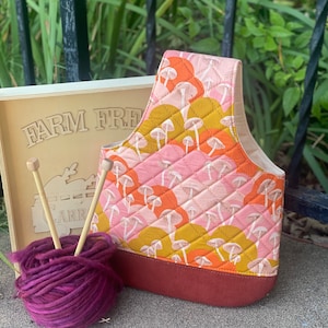 May include: A quilted tote bag with a pink, orange, and yellow mushroom pattern. The bag has a red bottom and is sitting on a concrete surface with a ball of purple yarn and two wooden knitting needles.