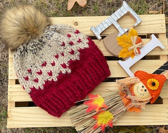 May include: A red and white knit hat with a brown faux fur pom pom. The hat is sitting on a wooden crate with fall decorations, including a scarecrow and a wooden sign that says "Fall".