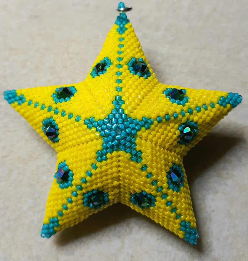 May include: A handmade, beaded star ornament in bright yellow and teal. The star is covered in small yellow beads with teal accents and iridescent gems. A teal star design is in the center. A small silver loop is at the top for hanging.