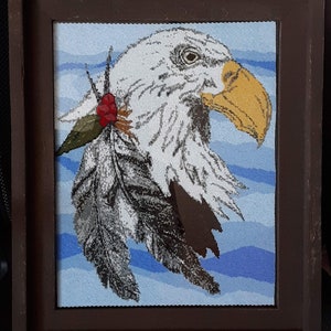 May include: A framed beaded artwork depicting a bald eagle with a red berry and green leaf on its head. The eagle is facing right and has a yellow beak. The background is a blue and white wavy pattern.