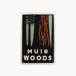 May include: A rectangular embroidered patch with a black background, featuring a stylized green tree and orange bark texture. The text MUIR WOODS is stitched in white capital letters at the bottom, all bordered by a white stitched edge.
