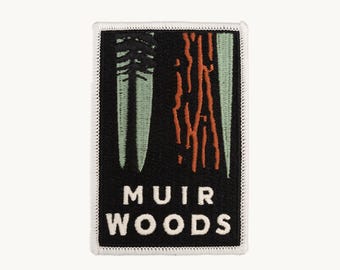 May include: A rectangular embroidered patch with a black background, featuring a stylized green tree and orange bark texture. The text MUIR WOODS is stitched in white capital letters at the bottom, all bordered by a white stitched edge.