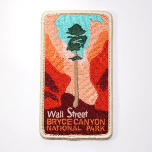 May include: A beige embroidered patch with a red, orange, and brown landscape scene. A green tree stands tall in the center of the patch. The text "Wall Street, Bryce Canyon National Park" is embroidered in white on the bottom of the patch.