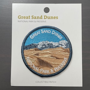 May include: A blue and white embroidered patch with the text "Great Sand Dunes National Park & Preserve".