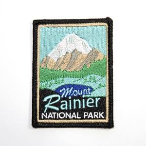 May include: An embroidered patch featuring a mountain scene with a blue sky, green trees, and a snow-capped peak. The text "Mount Rainier National Park" is embroidered in blue and white.
