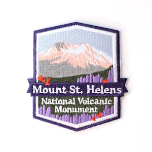May include: Embroidered patch featuring Mount St. Helens. The design includes a mountain with pink and gray tones, purple trees, and the text "Mount St. Helens National Volcanic Monument" in white lettering on a purple banner.