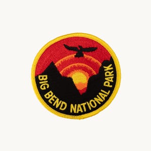 May include: A circular embroidered patch featuring a black silhouette of mountains and a bird against a red, orange, and yellow sunset. The text BIG BEND NATIONAL PARK is stitched in yellow along the bottom edge, bordered by a bright yellow rim.