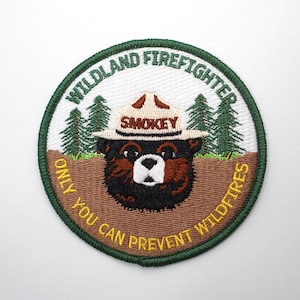 May include: A green and brown embroidered patch with a cartoon bear wearing a Smokey Bear hat. The text on the patch reads "Wildland Firefighter Only You Can Prevent Wildfires".