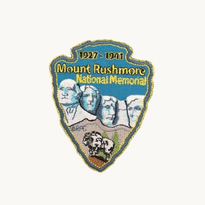 May include: A shield-shaped embroidered patch featuring the Mount Rushmore National Memorial with the text 1927-1941, Mount Rushmore, and National Memorial. The design includes the four presidential faces, a mountain goat, and trees.