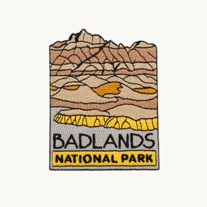 May include: An embroidered patch featuring a landscape of the Badlands National Park with layered rock formations in shades of tan, brown, and yellow. The text BADLANDS NATIONAL PARK is stitched in black lettering on a light gray and yellow background.