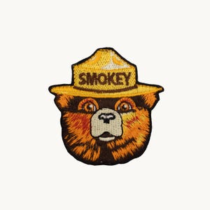 May include: An embroidered patch featuring the head of a bear wearing a yellow ranger hat with the word SMOKEY printed in black letters. The bear has brown and orange fur, light brown eyes, and a cream-colored muzzle with a black nose.