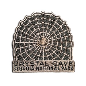 May include: A black and white embroidered patch with a spider web and a spider in the center. The text "CRYSTAL CAVE SEQUOIA NATIONAL PARK" is on the bottom of the patch.