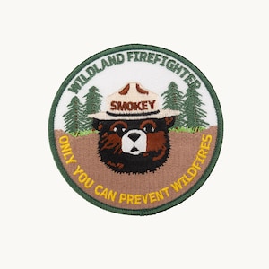 May include: A circular embroidered patch featuring a brown bear wearing a tan hat with the text SMOKEY. The background shows green pine trees and brown earth. The text WILDLAND FIREFIGHTER and ONLY YOU CAN PREVENT WILDFIRES is stitched.