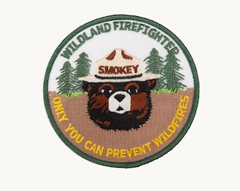 May include: A circular embroidered patch featuring the head of Smokey Bear wearing a tan hat with the word SMOKEY. The background shows green pine trees and brown earth. The text WILDLAND FIREFIGHTER and ONLY YOU CAN PREVENT WILDFIRES is stitched.