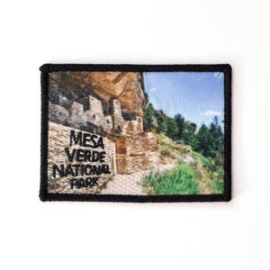 May include: A black and white embroidered patch with a photo of Mesa Verde National Park. The text "Mesa Verde National Park" is embroidered in black on the patch.