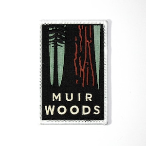 May include: A black and white embroidered patch with the text "MUIR WOODS" in white. The patch features a stylized image of two trees with green leaves and red bark.