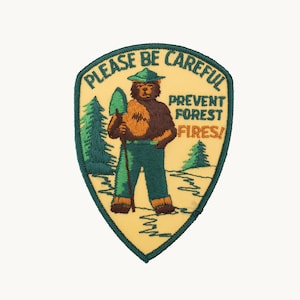 May include: A shield-shaped embroidered patch with a cream background and dark green border. It features a brown bear wearing a green hat and pants, holding a shovel. The text reads: PLEASE BE CAREFUL PREVENT FOREST FIRES! with green pine trees.