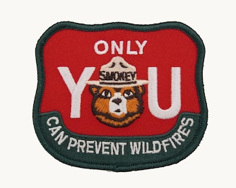 Może przedstawiać: Wyszywana naszywka w kształcie tarczy z czerwonym tłem i ciemnozieloną obwódką. Zawiera biały napis ONLY YOU oraz CAN PREVENT WILDFIRES, a także centralną ilustrację niedźwiedzia Smokey Bear w beżowym kapeluszu strażnika.