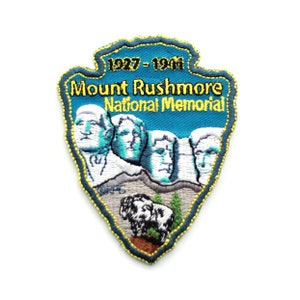 May include: A blue and yellow embroidered patch with the text "Mount Rushmore National Memorial" and the dates "1927 - 1941". The patch features a depiction of the four presidents carved into the mountain, with a mountain goat in the foreground.