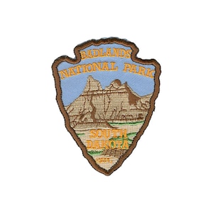 May include: Embroidered arrowhead-shaped patch with the text "BADLANDS NATIONAL PARK SOUTH DAKOTA". The patch shows a landscape with mountains, a blue sky, and green grass, framed by a brown border.