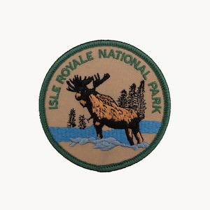 May include: A circular embroidered patch featuring a tan background with a dark green border. The text ISLE ROYALE NATIONAL PARK is arched in green. A brown moose stands in a blue river with grey rocks and black pine trees in the background.
