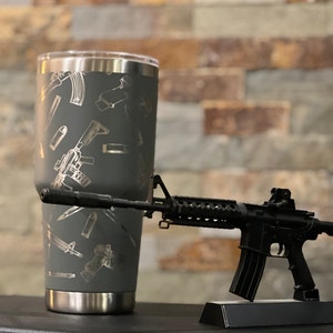 May include: A gray stainless steel tumbler with a silver lid. The tumbler is covered in a pattern of silver-colored guns and bullets. A black toy rifle is in the background.