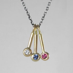 Puede incluir: Un collar de oro con tres colgantes, cada uno con una gema de color diferente: un diamante blanco, un zafiro azul y un rubí rojo.