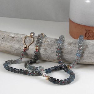 Puede incluir: Un collar largo hecho de cuentas de piedras preciosas azules y grises con detalles dorados y un cierre dorado. El collar se muestra sobre un trozo de madera a la deriva.