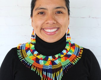 May include: A woman wearing a black turtleneck sweater with a colourful beaded necklace and matching earrings. The necklace is a large, geometric design with fringe. The earrings are long and dangle. The woman is smiling and looking at the camera. The background is a white brick wall. @latinxculturviva