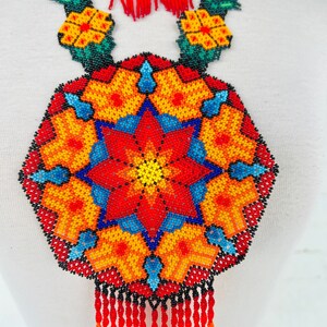 May include: A colorful beaded necklace with a large, round pendant featuring a starburst design in shades of red, orange, yellow, blue, and green. The pendant is surrounded by smaller floral motifs and the necklace has a fringe of red beads.