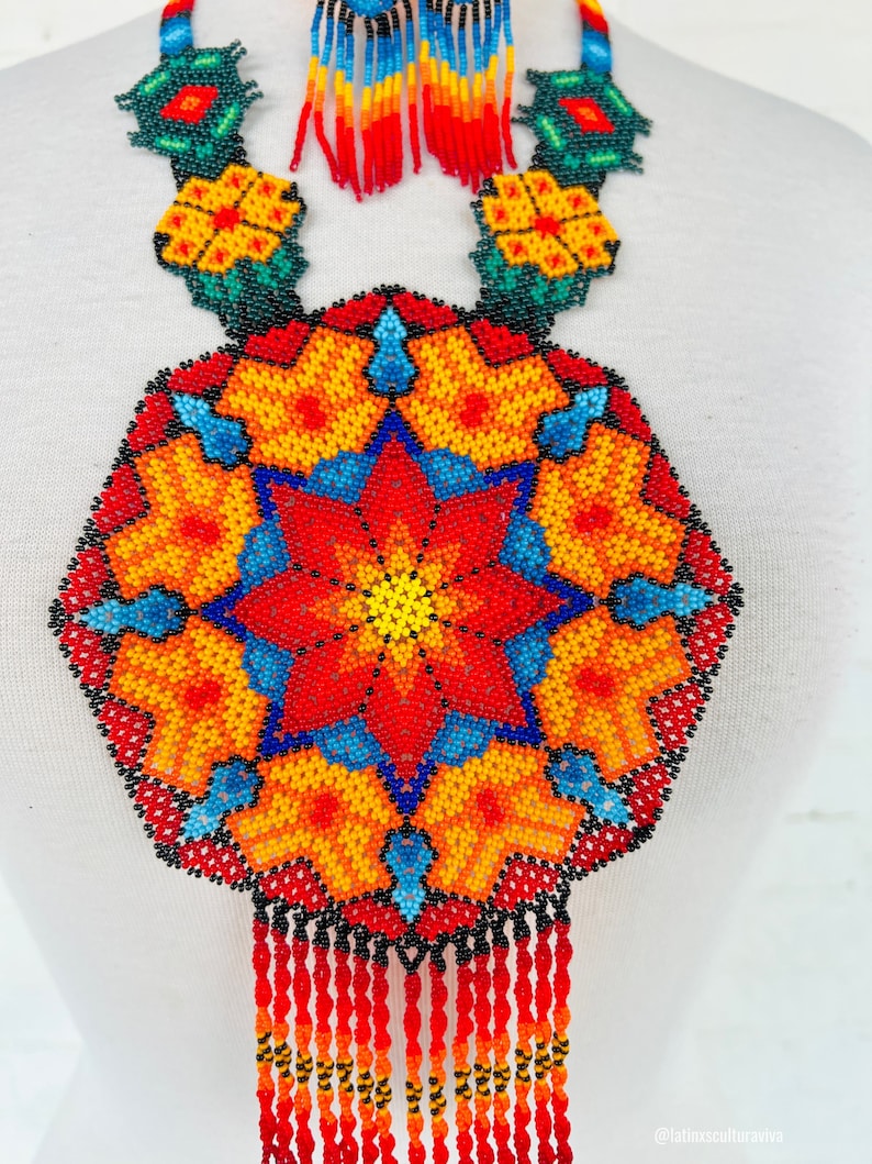 May include: A colorful beaded necklace with a large, round pendant featuring a starburst design in shades of red, orange, yellow, blue, and green. The pendant is surrounded by smaller floral motifs and the necklace has a fringe of red beads.