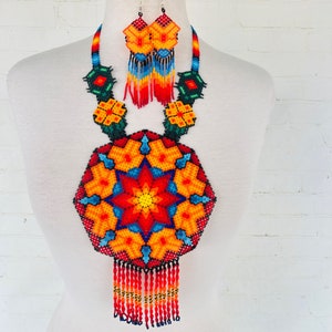May include: A colorful beaded necklace and earrings set with a large, round pendant featuring a starburst design in shades of red, orange, yellow, blue, and green. The necklace has a fringe of beads.