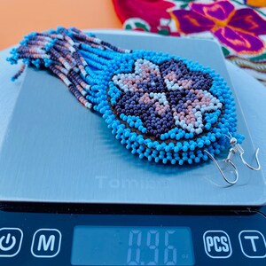 K&ouml;nnte beinhalten: Ein Paar Perlenohrringe mit einem blauen, wei&szlig;en, rosa und violetten Blumenmuster. Die Ohrringe befinden sich auf einer digitalen Waage, die 0,95 oz anzeigt. Der Text "@latinxsculturaviva" befindet sich auf der Waage.