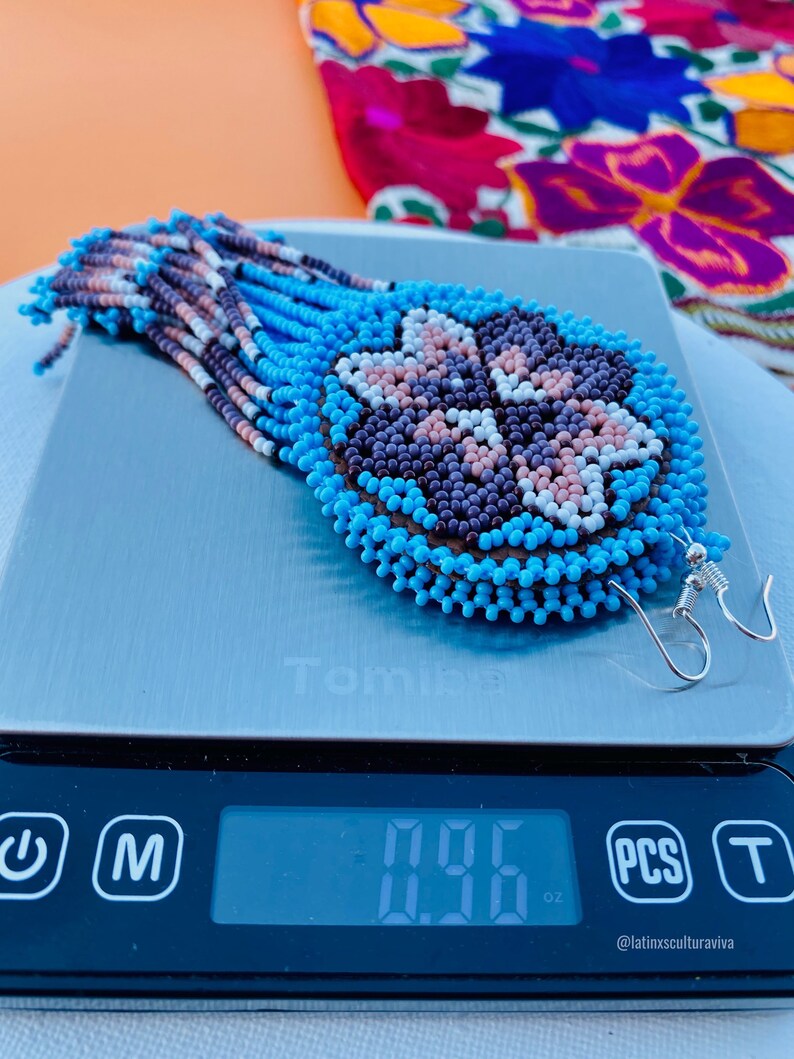 K&ouml;nnte beinhalten: Ein Paar Perlenohrringe mit einem blauen, wei&szlig;en, rosa und violetten Blumenmuster. Die Ohrringe befinden sich auf einer digitalen Waage, die 0,95 oz anzeigt. Der Text "@latinxsculturaviva" befindet sich auf der Waage.