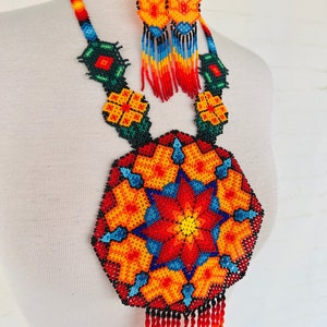 May include: A colorful beaded necklace and earrings set with a large, round pendant featuring a starburst design in red, orange, yellow, blue, and black. The pendant has a fringe of red beads.