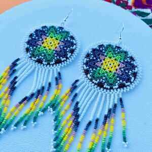 以下が含まれることがあります： 手作りのビーズイヤリング。中央に黄色の花、周囲に緑、青、茶色のビーズをあしらった円形デザイン。下には白いフリンジがあり、黄色、緑、青のカラフルなアクセントが施されています。