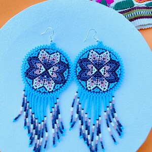 K&ouml;nnte beinhalten: Ein Paar blaue und wei&szlig;e Perlenohrringe mit einem Blumenmuster. Die Ohrringe sind aus kleinen, bunten Perlen gefertigt und haben einen Fransen aus Perlen, der von unten h&auml;ngt.