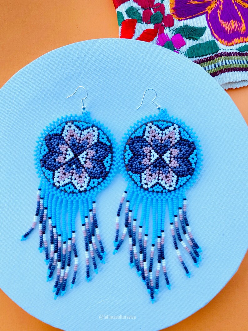 K&ouml;nnte beinhalten: Ein Paar blaue und wei&szlig;e Perlenohrringe mit einem Blumenmuster. Die Ohrringe sind aus kleinen, bunten Perlen gefertigt und haben einen Fransen aus Perlen, der von unten h&auml;ngt.