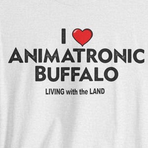 May include: A light gray t-shirt with the text "I ❤️ ANIMATRONIC BUFFALO" in black, with a red heart symbol. Below the text is the phrase "LIVING with the LAND" in a smaller font. The shirt has short sleeves and a crew neck.