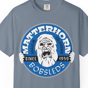 May include: A blue-grey t-shirt featuring a circular graphic with a white yeti-like creature with red eyes. The graphic reads "Matterhorn Anaheim California Since 1959 Bobsleds" in white and blue.