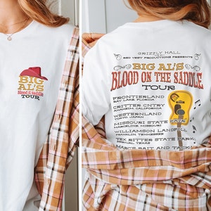 Könnte beinhalten: Weißes T-Shirt mit einer roten und gelben Grafik eines Cowboyhuts und dem Text "Big Al's Blood on the Saddle Tour". Auf der Rückseite des Shirts ist eine Liste der Tourdaten und -orte.