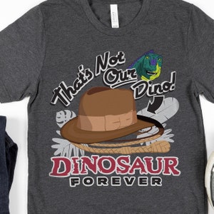 Op de afbeelding: Een donkergrijs T-shirt met een cartoon dinosaurus, een fedora-hoed en een zweep. De tekst op het shirt luidt "That's Not Our Dino! Dinosaur Forever".